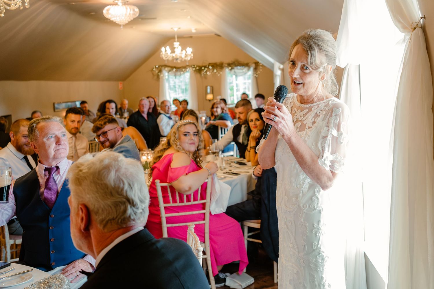 A wedding reception scene with guests seated at tables while someone gives a speech.