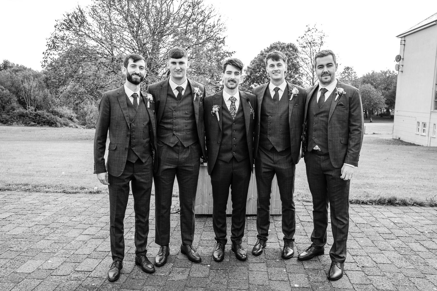Groomsmen in dark suits stand together outdoors for formal wedding photos.