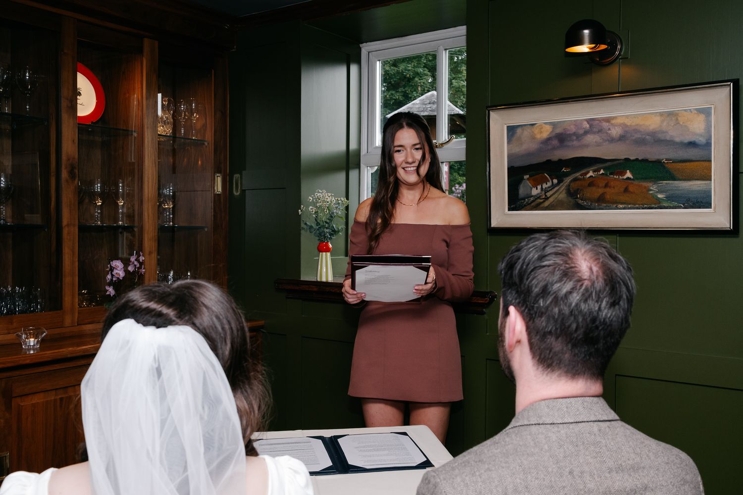 Woman in brown dress reads at intimate wedding ceremony in green-walled venue with guests seated before her.