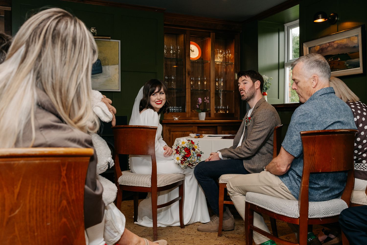 Bride and groom sit with guests in a cozy green-walled venue, bride holding a colorful bouquet during wedding celebration.