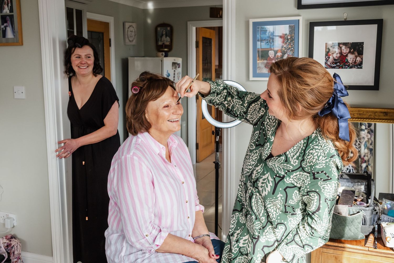 A woman gets her hair styled while two friends watch and smile in a cozy home setting.
