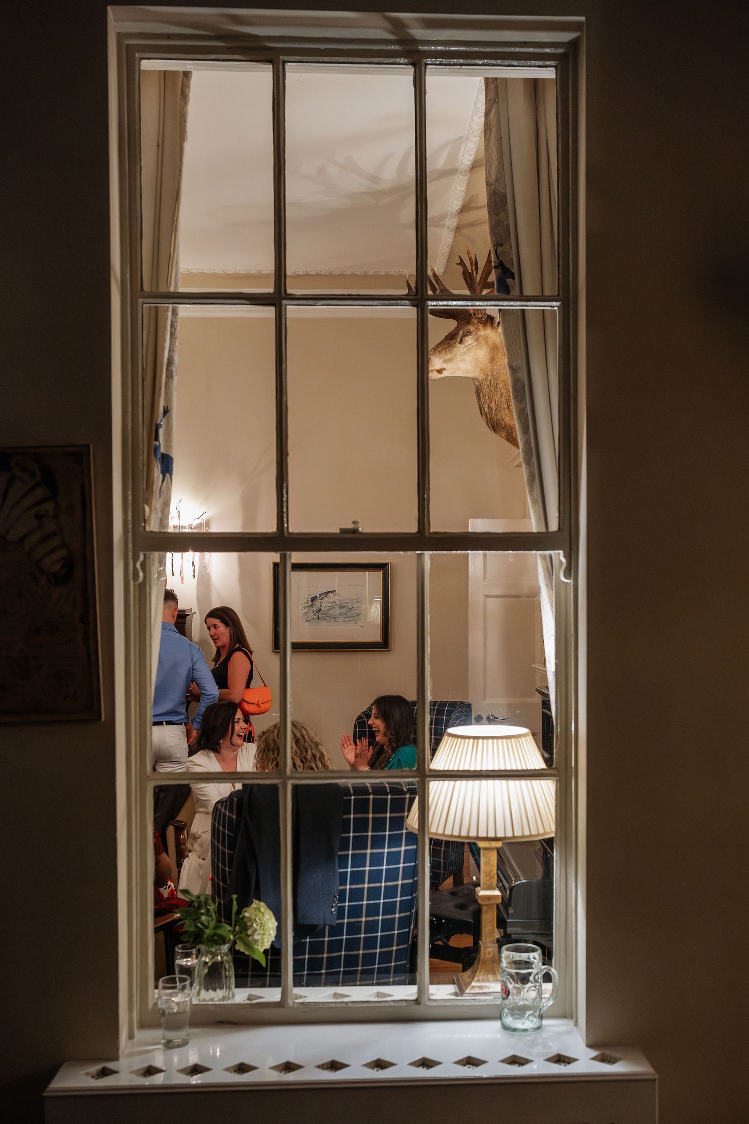 Wedding guests socializing inside a venue viewed through a multi-pane window with lamp.