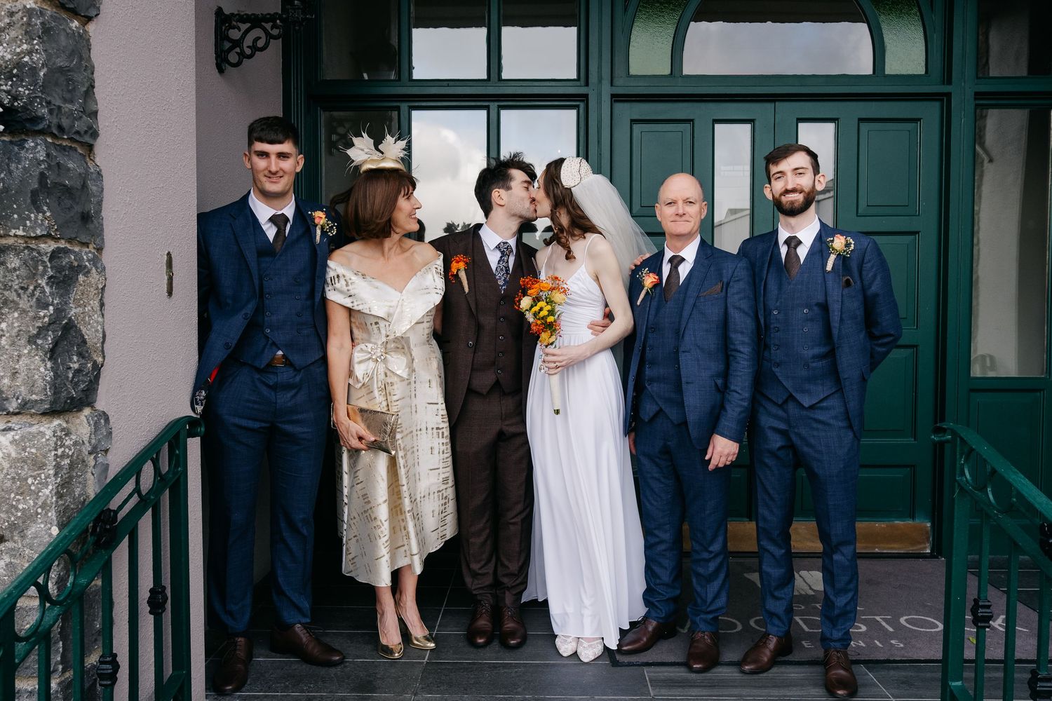 Wedding party members pose together in formal attire in front of a dark green door.