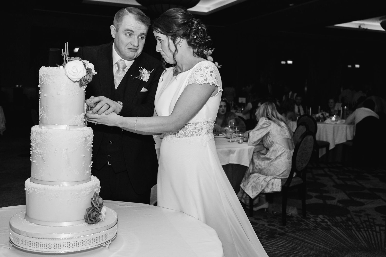 Elegant white wedding cake displayed at reception with couples dancing and celebrating in the background.