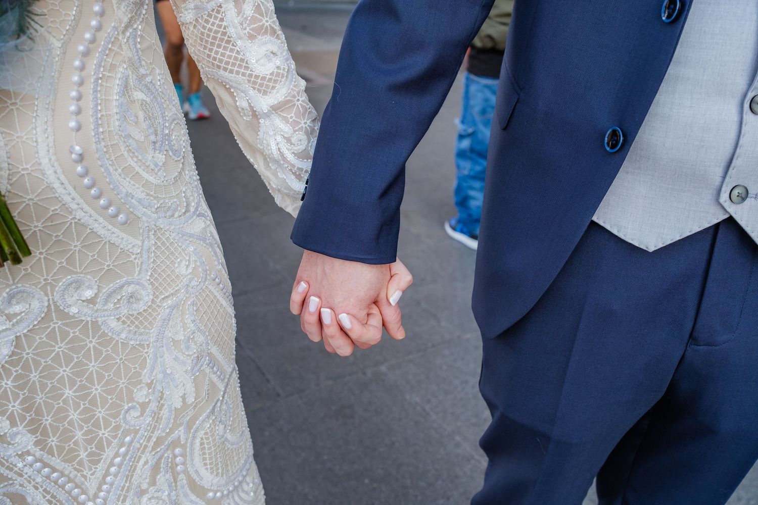 Close-up of newlywed couple holding hands, showing lace dress and navy suit details.