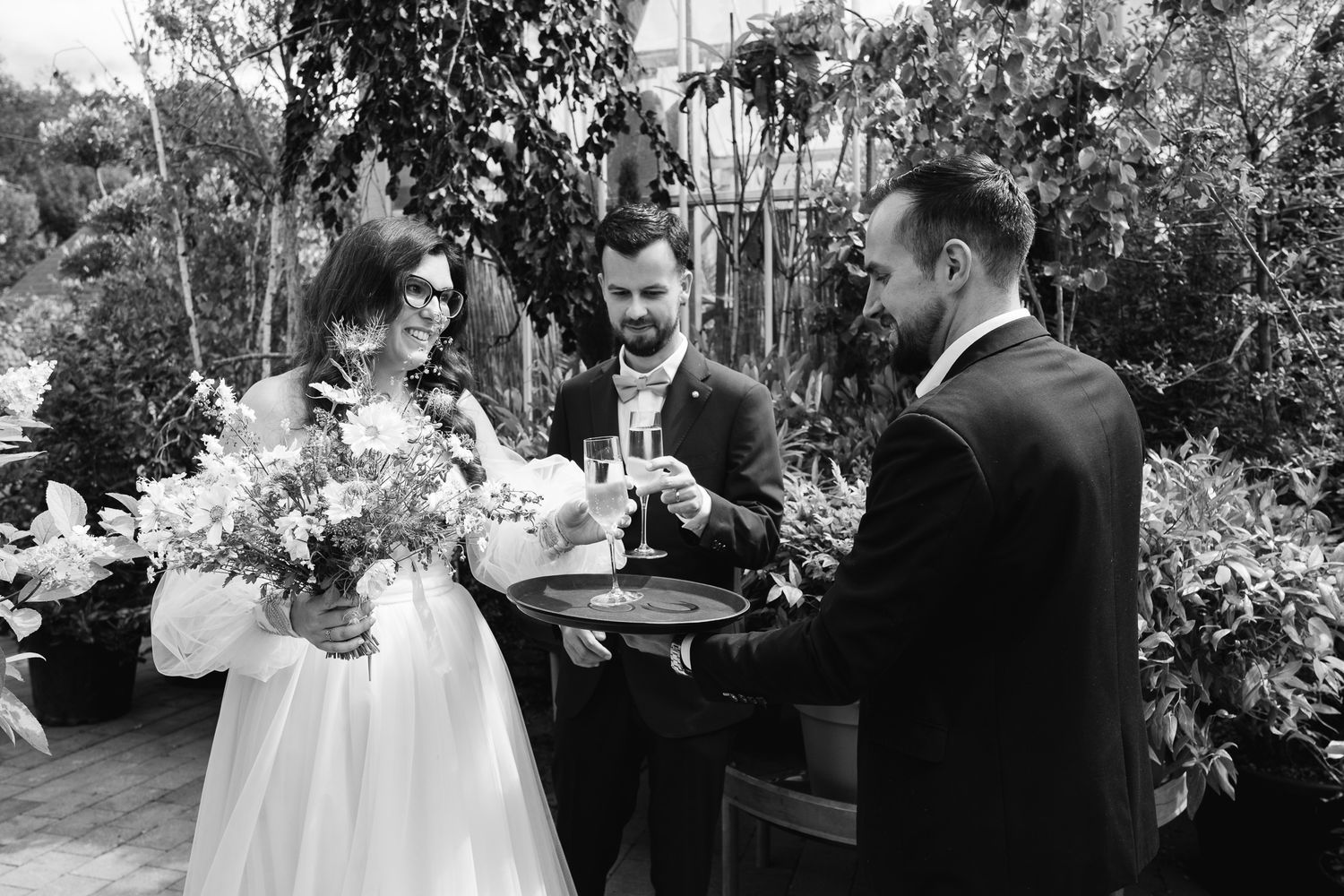 Black and white wedding photo of bride and groom receiving drinks on a tray outdoors.