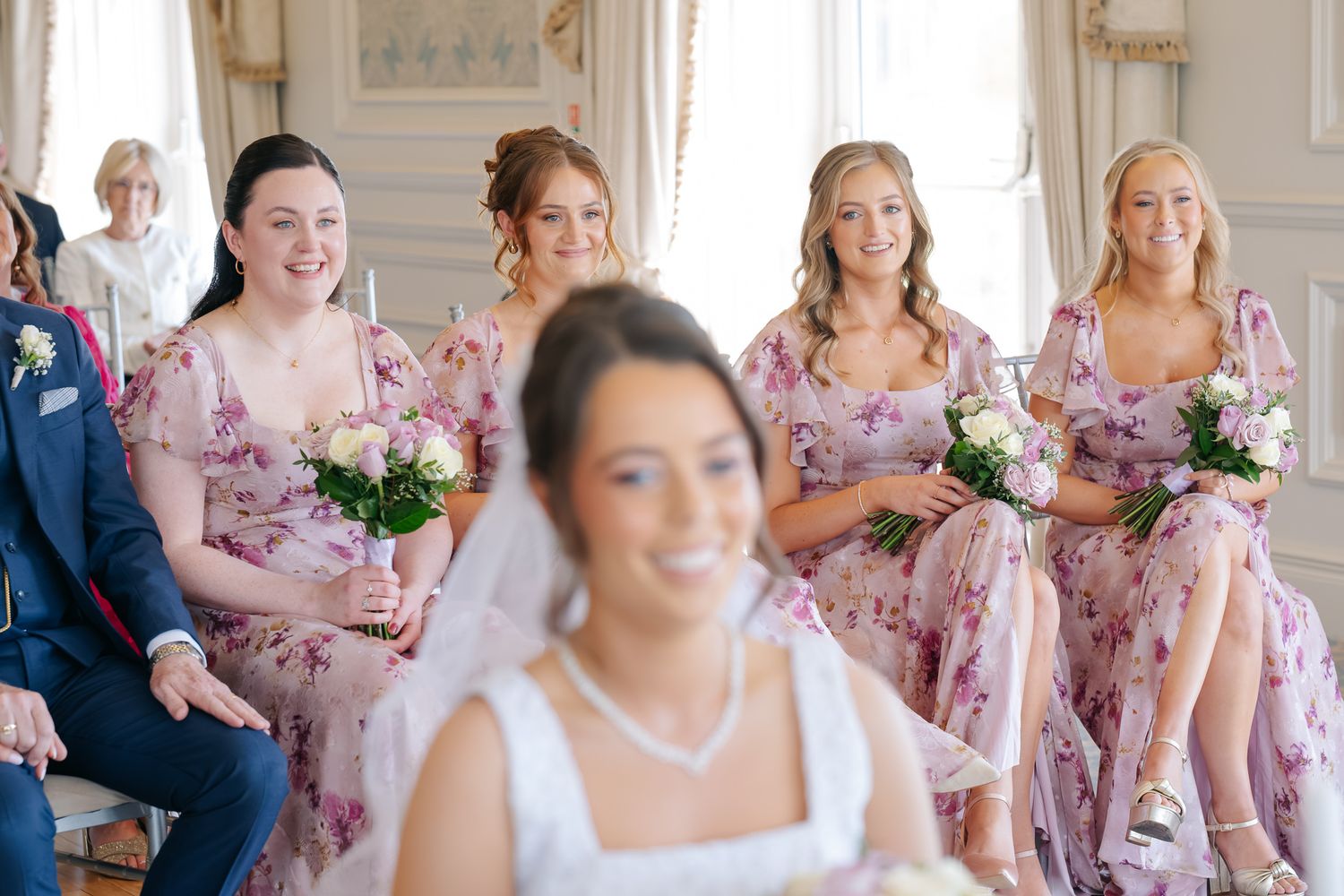 Bride in white veil smiles as bridesmaids in pink floral dresses hold bouquets at an elegant wedding ceremony.