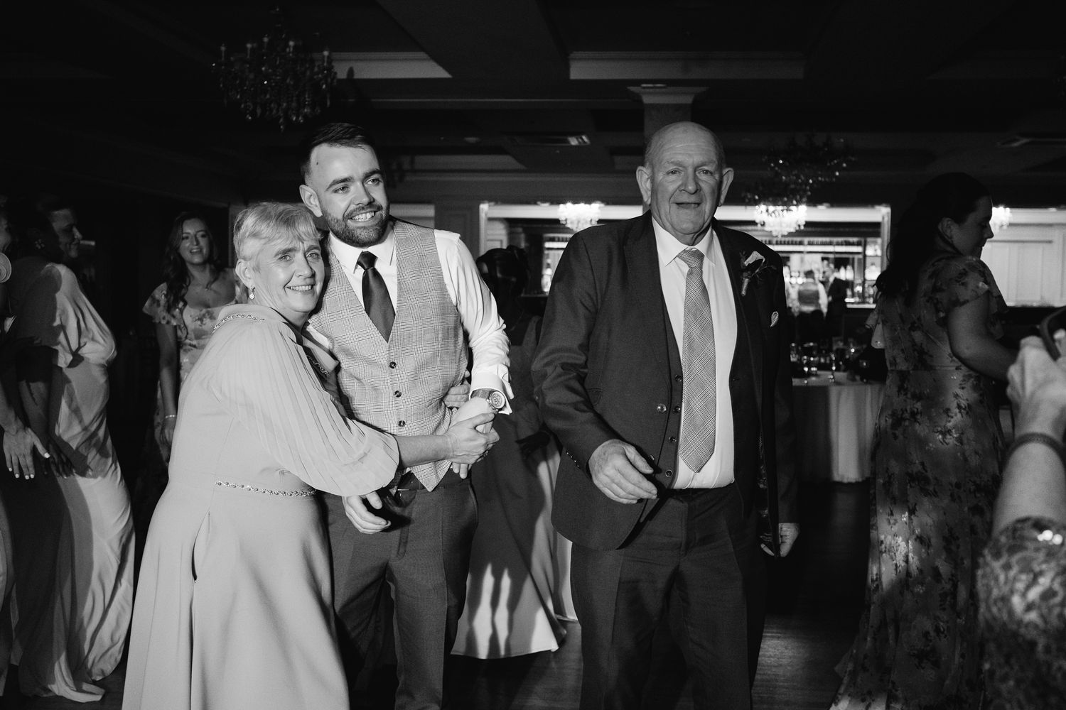 Black and white wedding reception photo of guests dancing and smiling joyfully at an elegant venue.