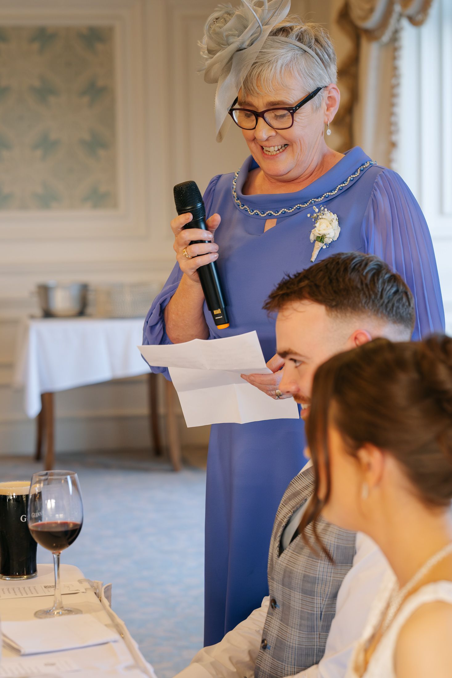 Older woman in blue dress giving a speech with microphone at a wedding reception while guests look on.