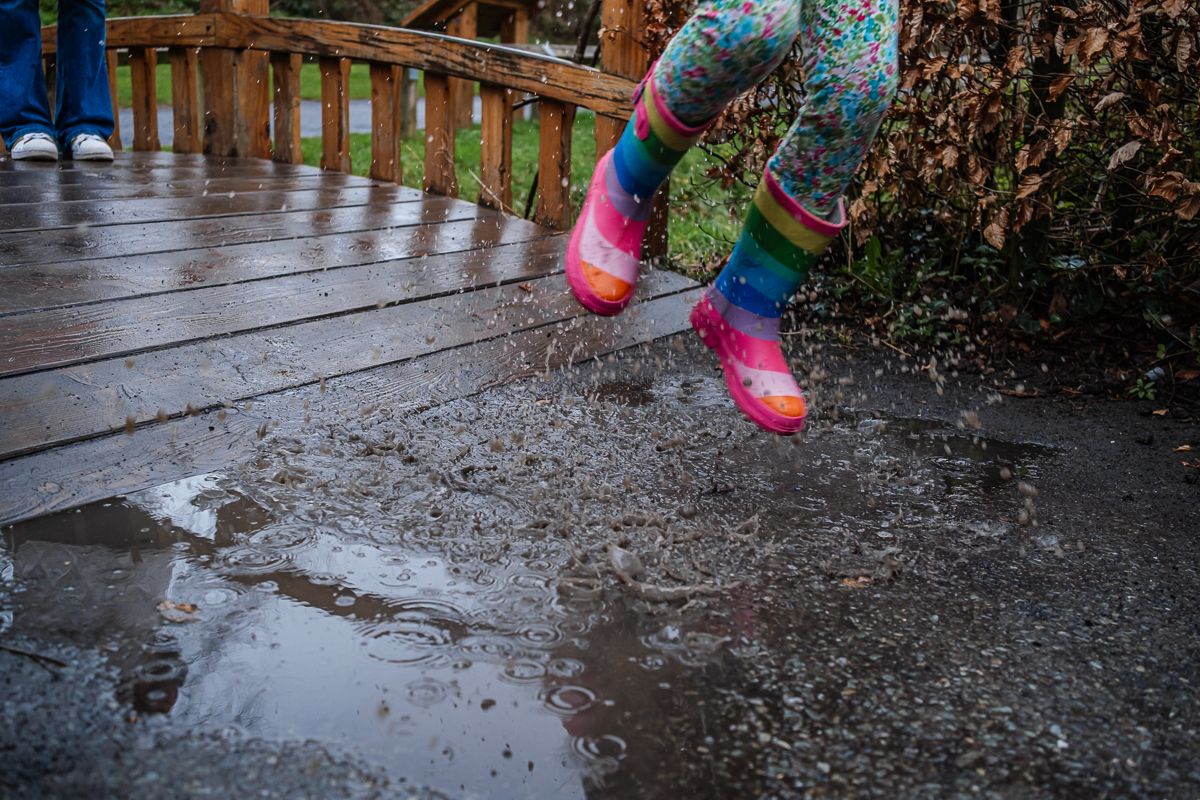 Child in colorful rainbow rain boots jumping in muddy puddle on wooden deck during rainy day.
