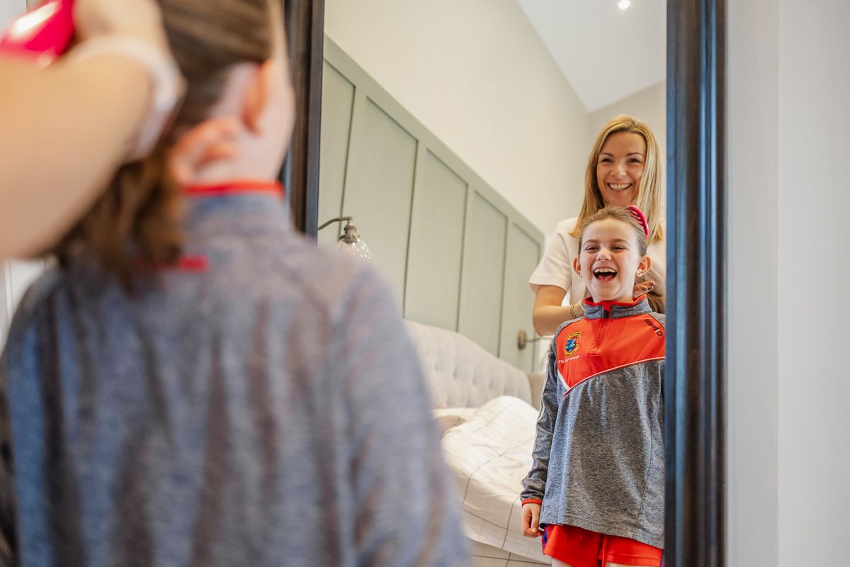 A mother helps her laughing child brush teeth in front of a mirror in a bright modern bathroom.
