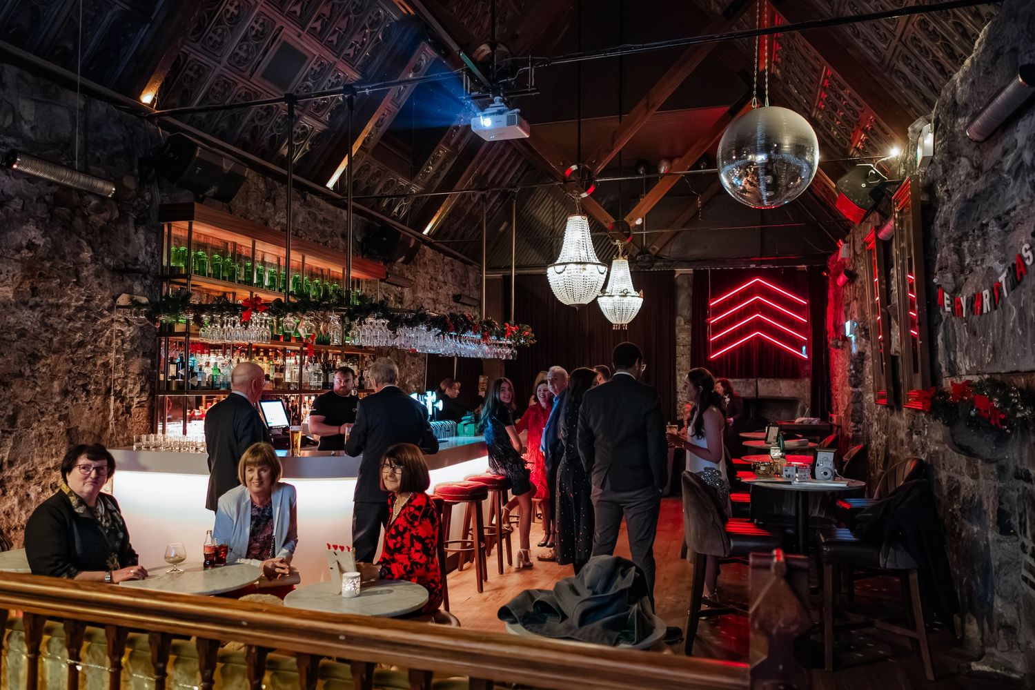 Dimly lit rustic bar with stone walls, disco ball, chandeliers, and patrons socializing near a glowing counter.