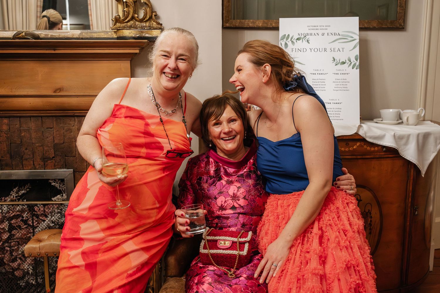 Three women in colorful formal dresses smiling and hugging at an elegant wedding reception venue.