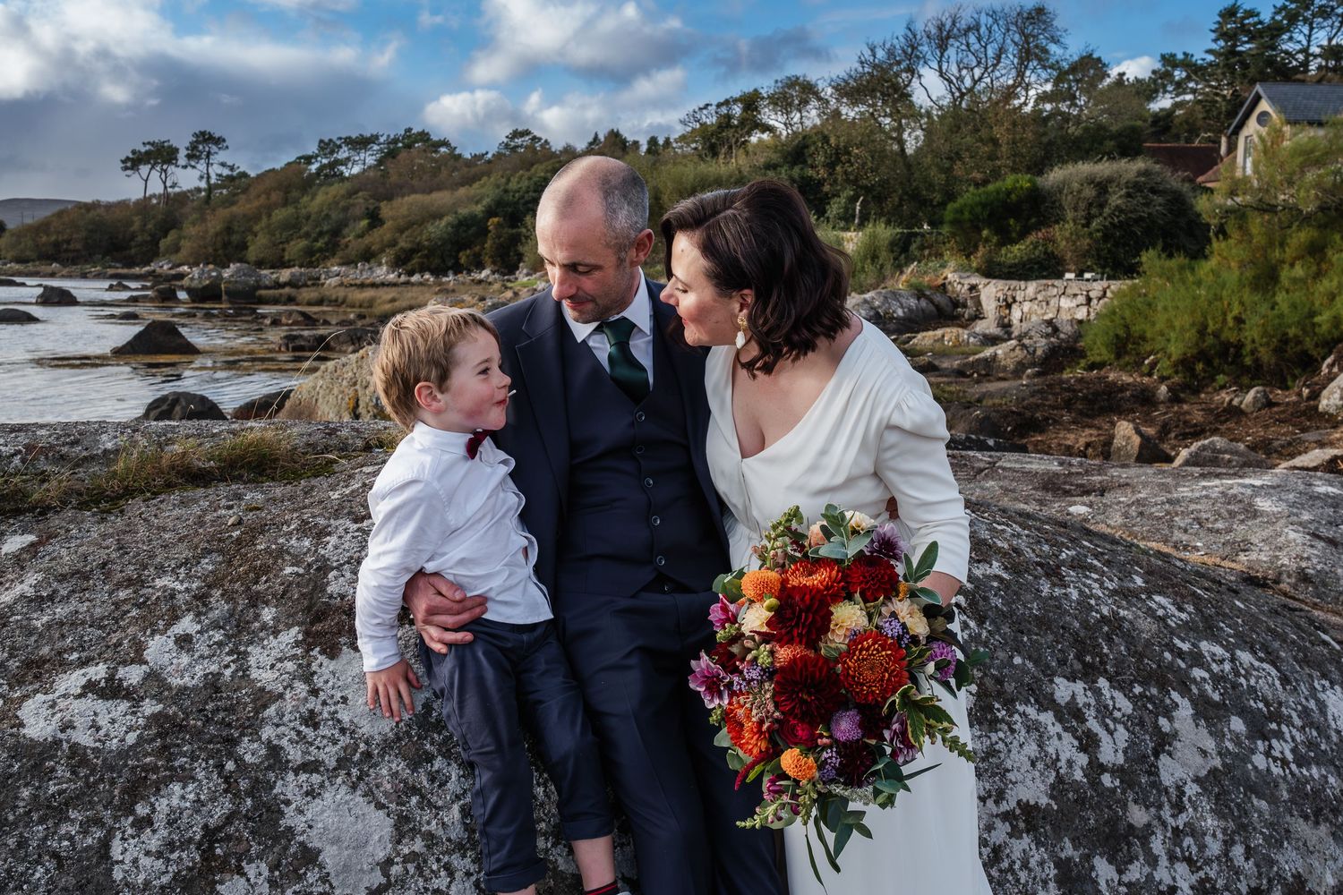 Bride and groom with young boy in red bow tie posing on coastal rocks with colorful bouquet on cloudy day.
