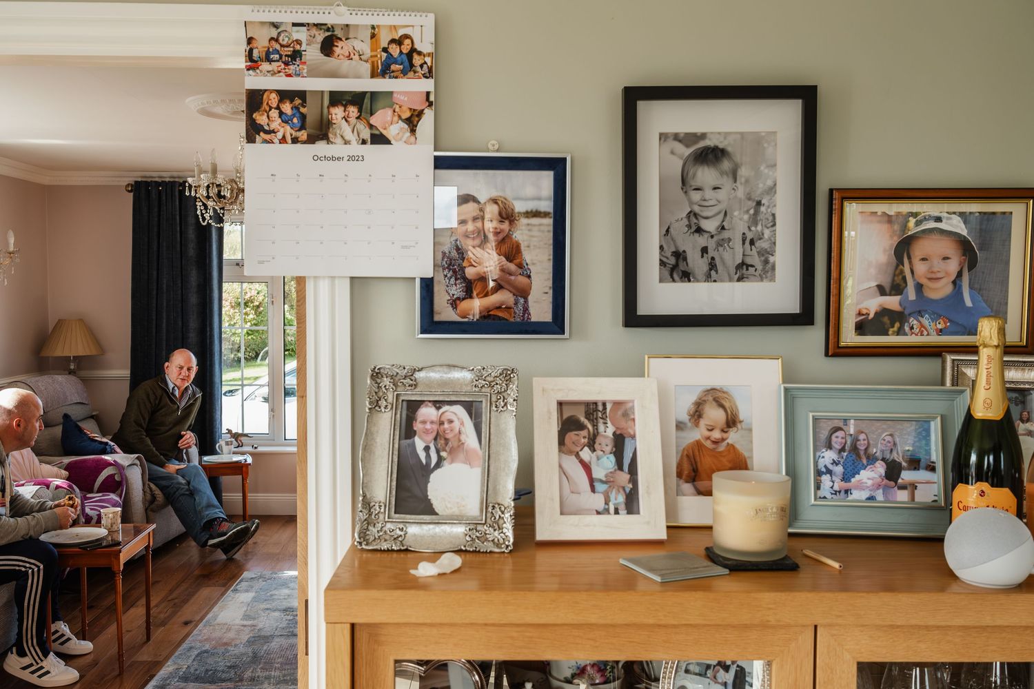 Family photo wall display with framed portraits and calendar above a wooden sideboard in a cozy living room.