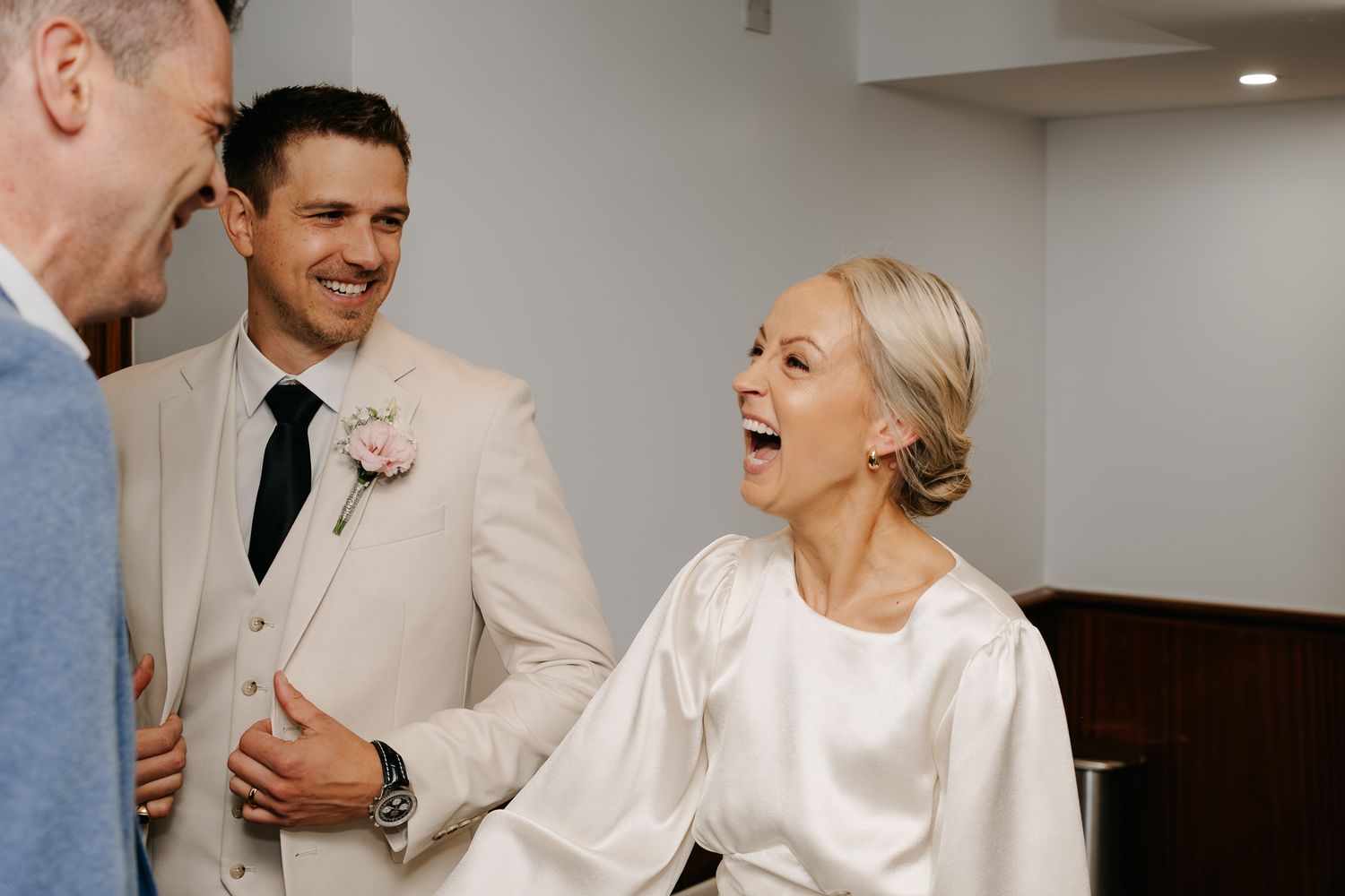 A bride in a white satin robe laughs joyfully with a groom in a beige suit during wedding preparations.