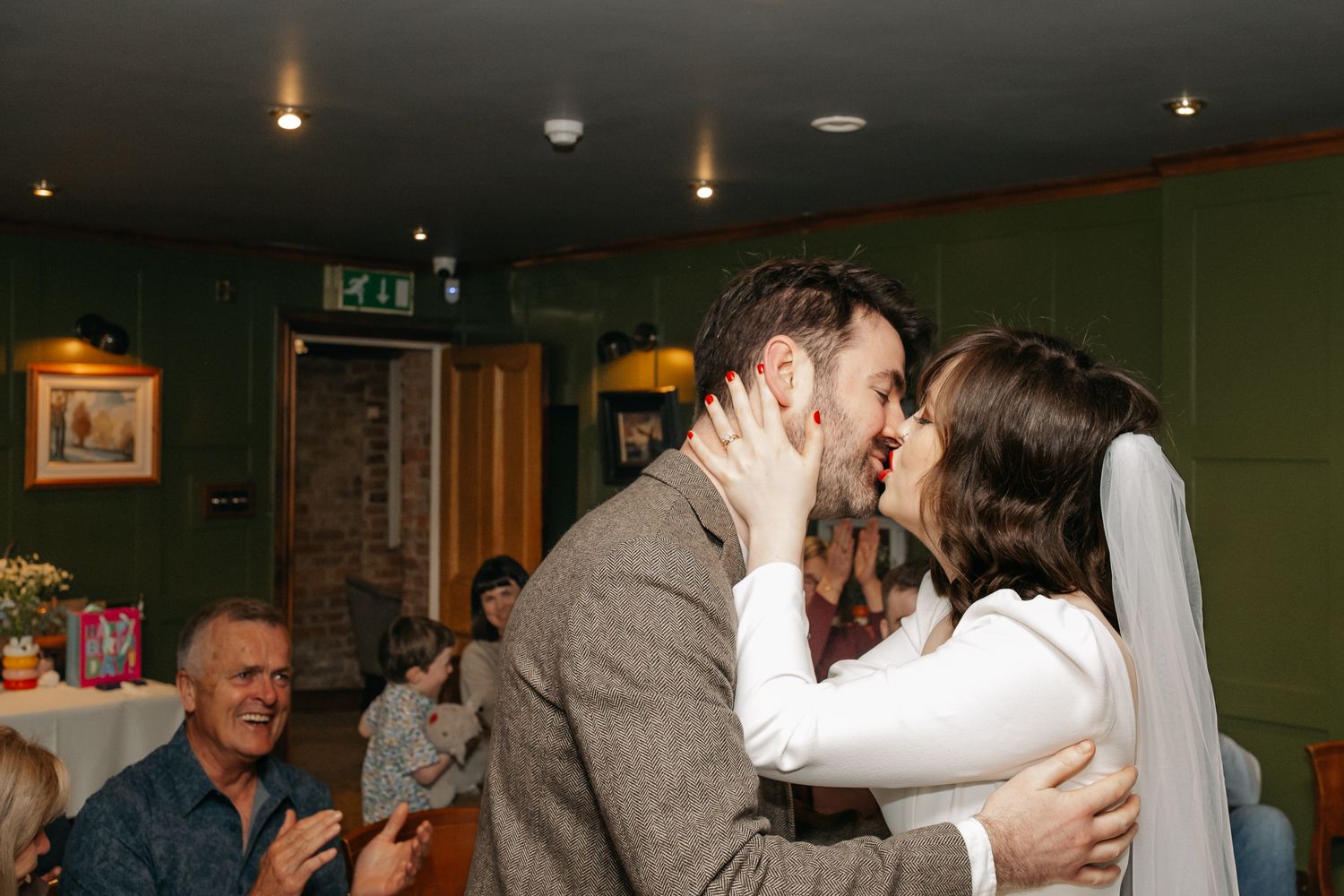 Bride and groom share a first kiss at wedding reception while guests applaud in a cozy green-walled venue.