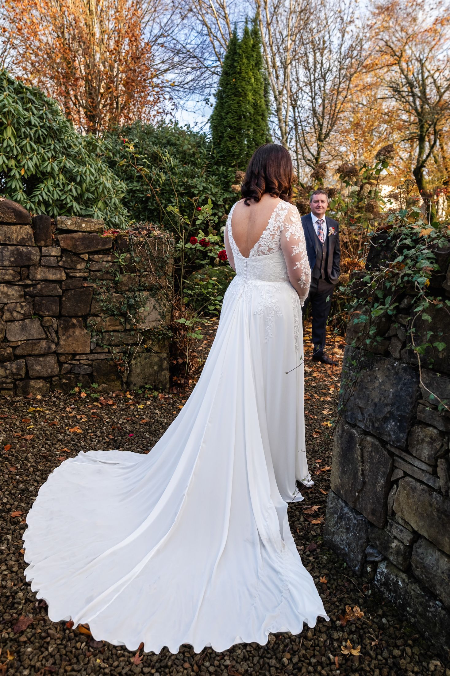 Bride in elegant white lace gown with long train stands in autumn garden as groom watches in background.