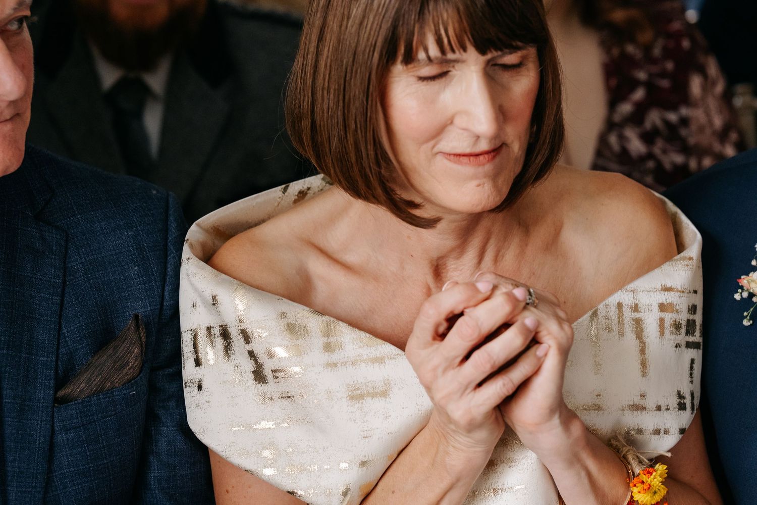 Woman in off-shoulder sequined dress clasping hands, eyes closed, at a wedding ceremony.