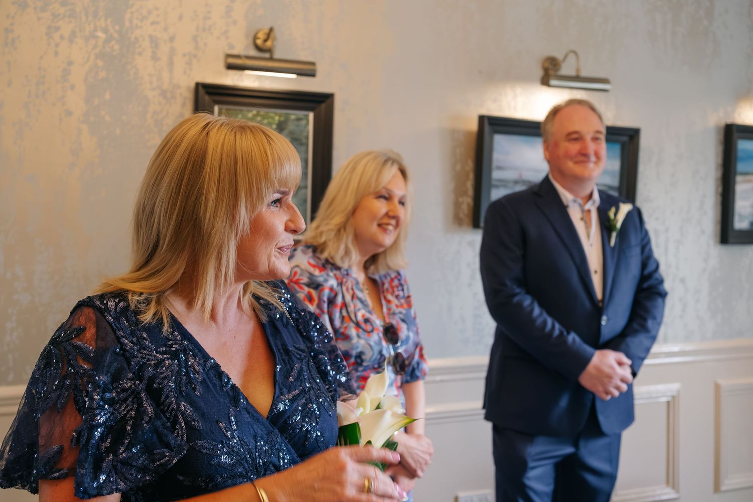 Two women in elegant dresses smile warmly as a man in a blue suit stands in the background at a wedding ceremony.