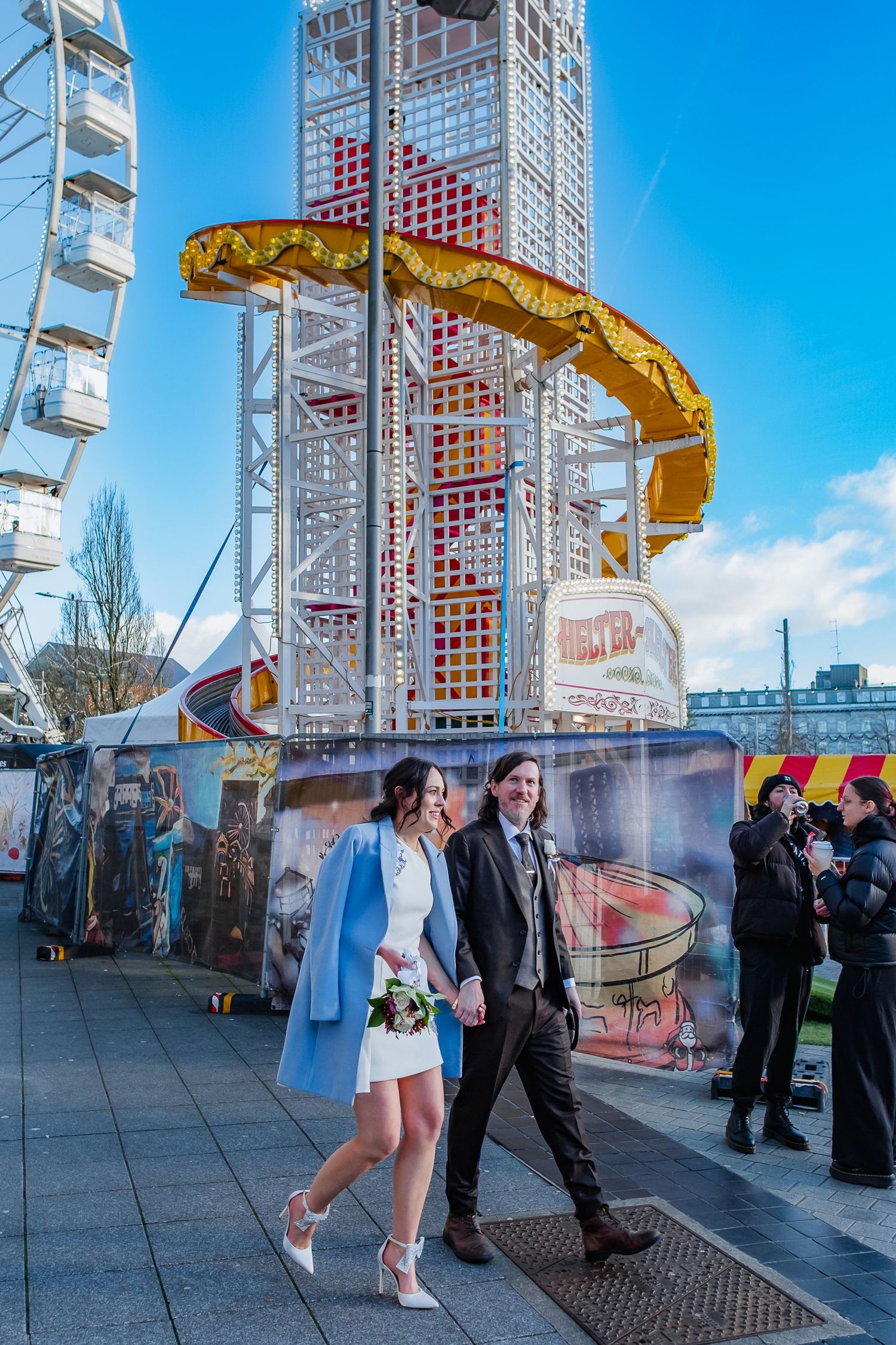 Bride and groom walking hand in hand at a fairground with a Ferris wheel and helter-skelter on a sunny day.