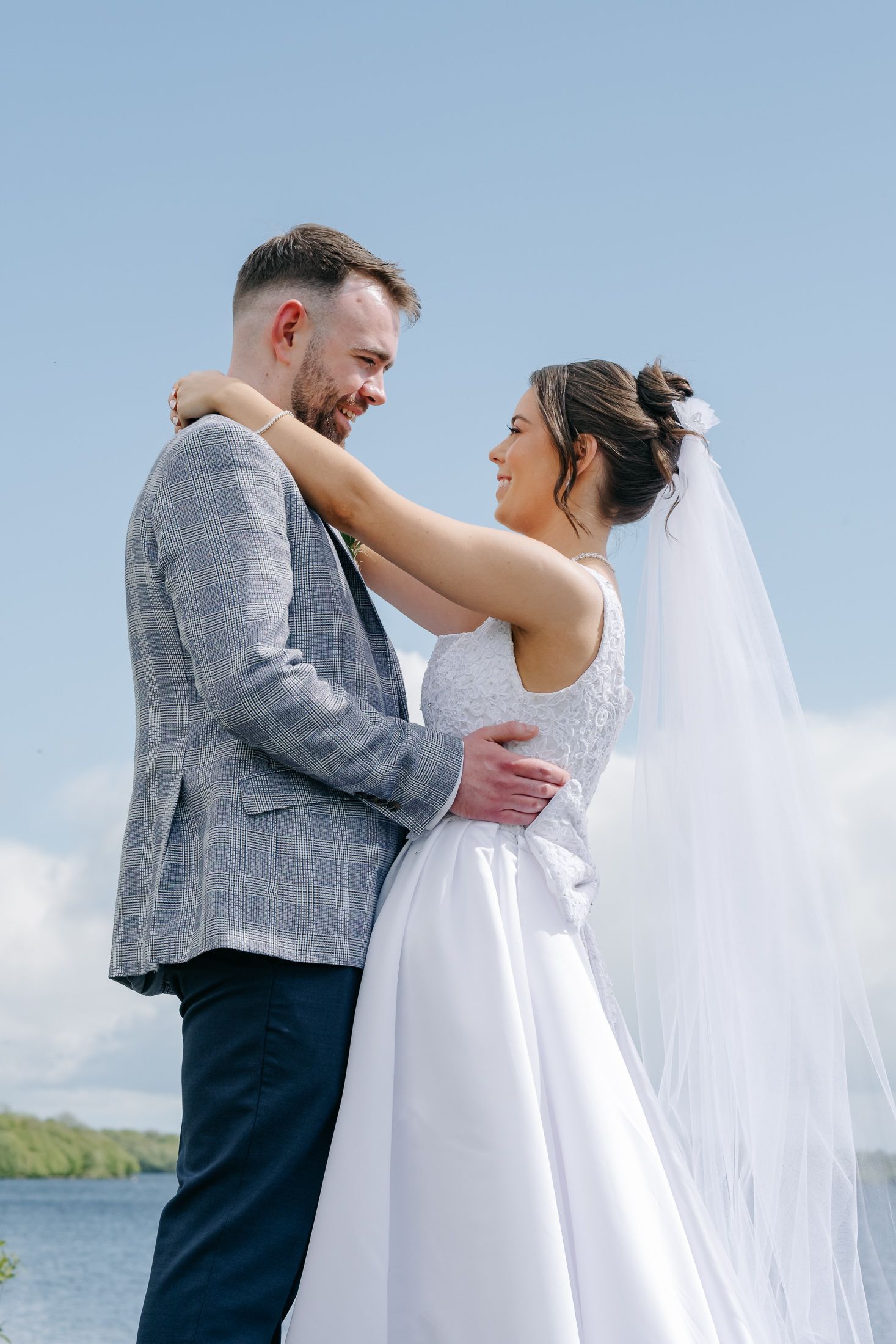 Bride and groom embrace outdoors by a lake, smiling at each other, bride in white gown with veil, groom in gray suit.