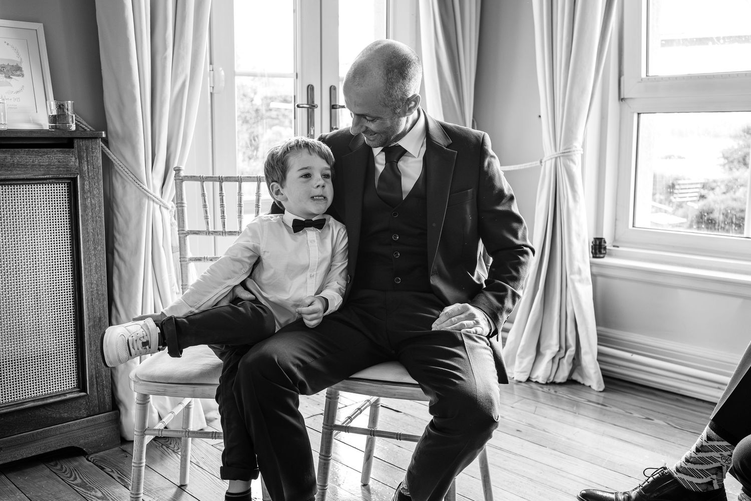 A man in a suit shares a tender moment with a young boy in a bow tie at a wedding.