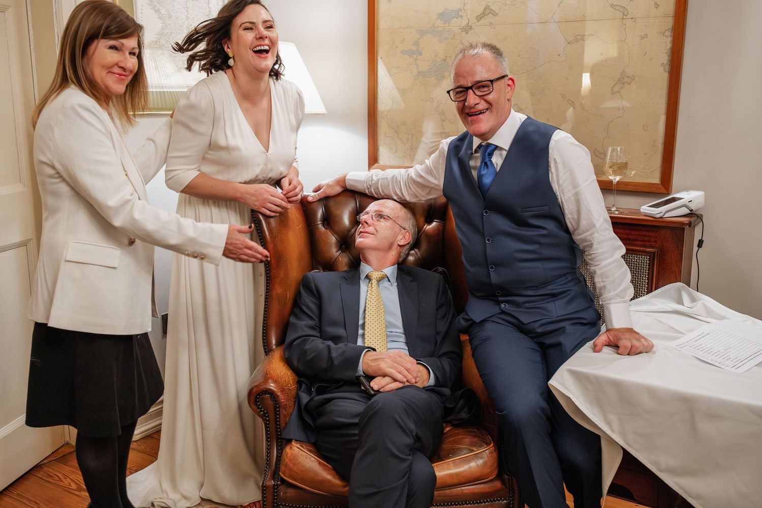 A joyful wedding group laughing together, bride in white dress with three guests in formal attire indoors.