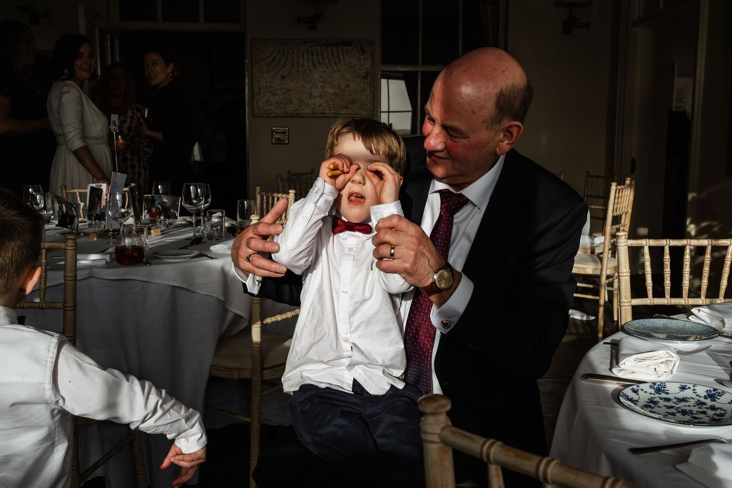 Man playfully covering a young boy's eyes at a wedding reception dinner table.