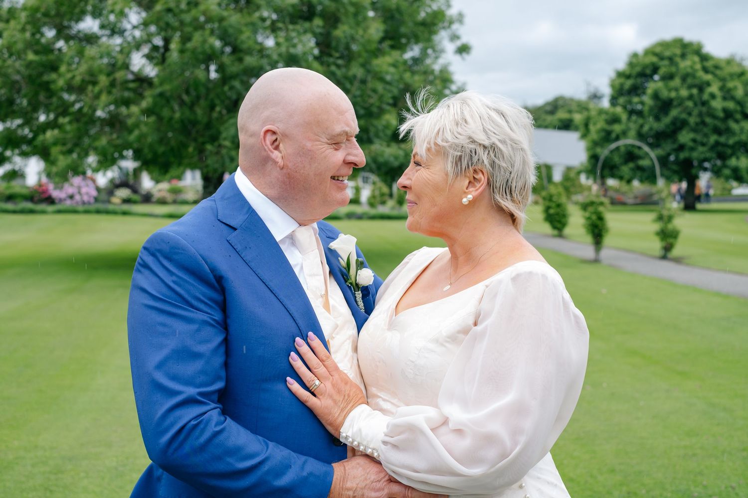 A couple in wedding attire smiling at each other on a green lawn, groom in blue suit, bride in white dress.