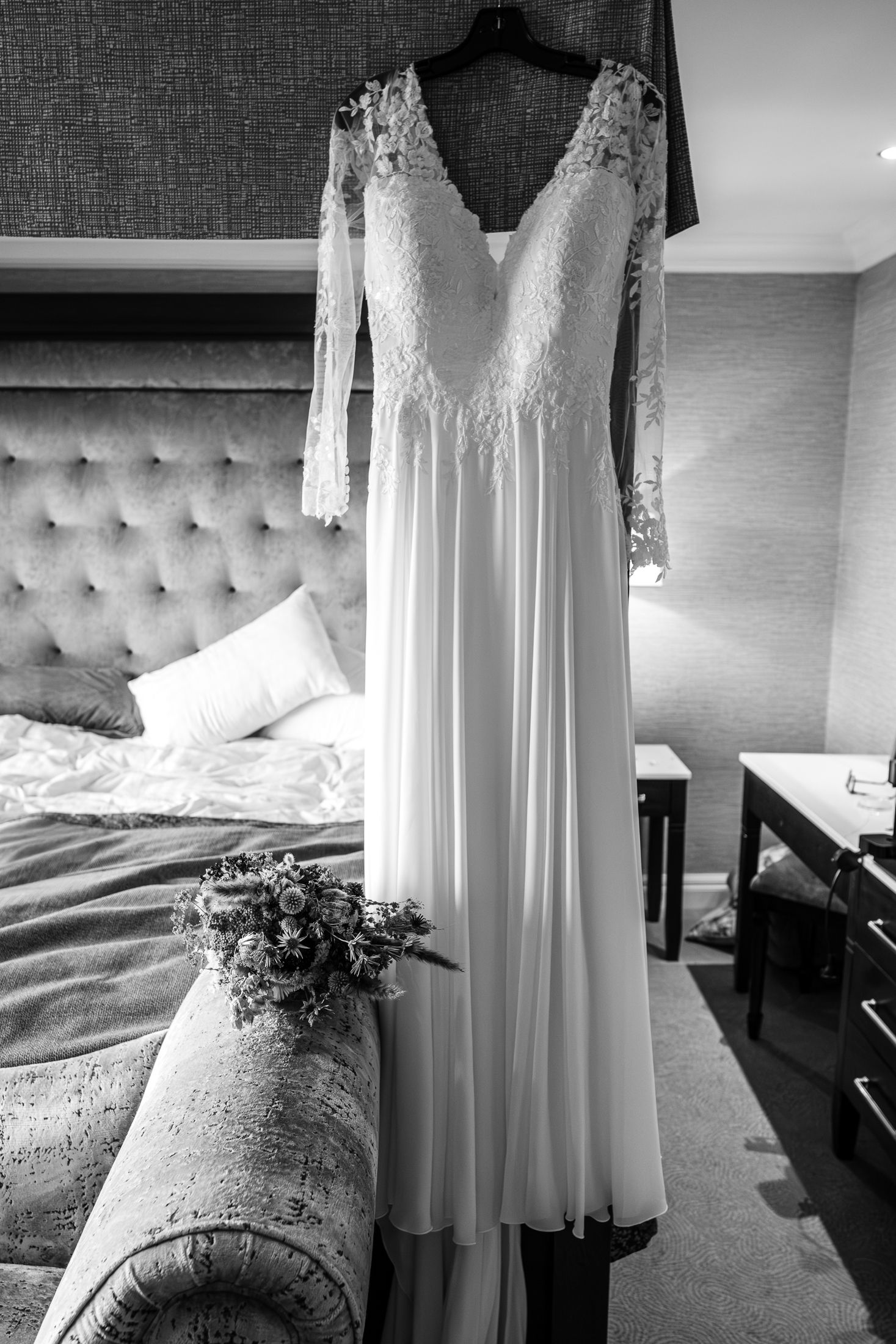 Elegant long-sleeve lace wedding dress hanging in hotel bedroom with bridal bouquet in black and white.