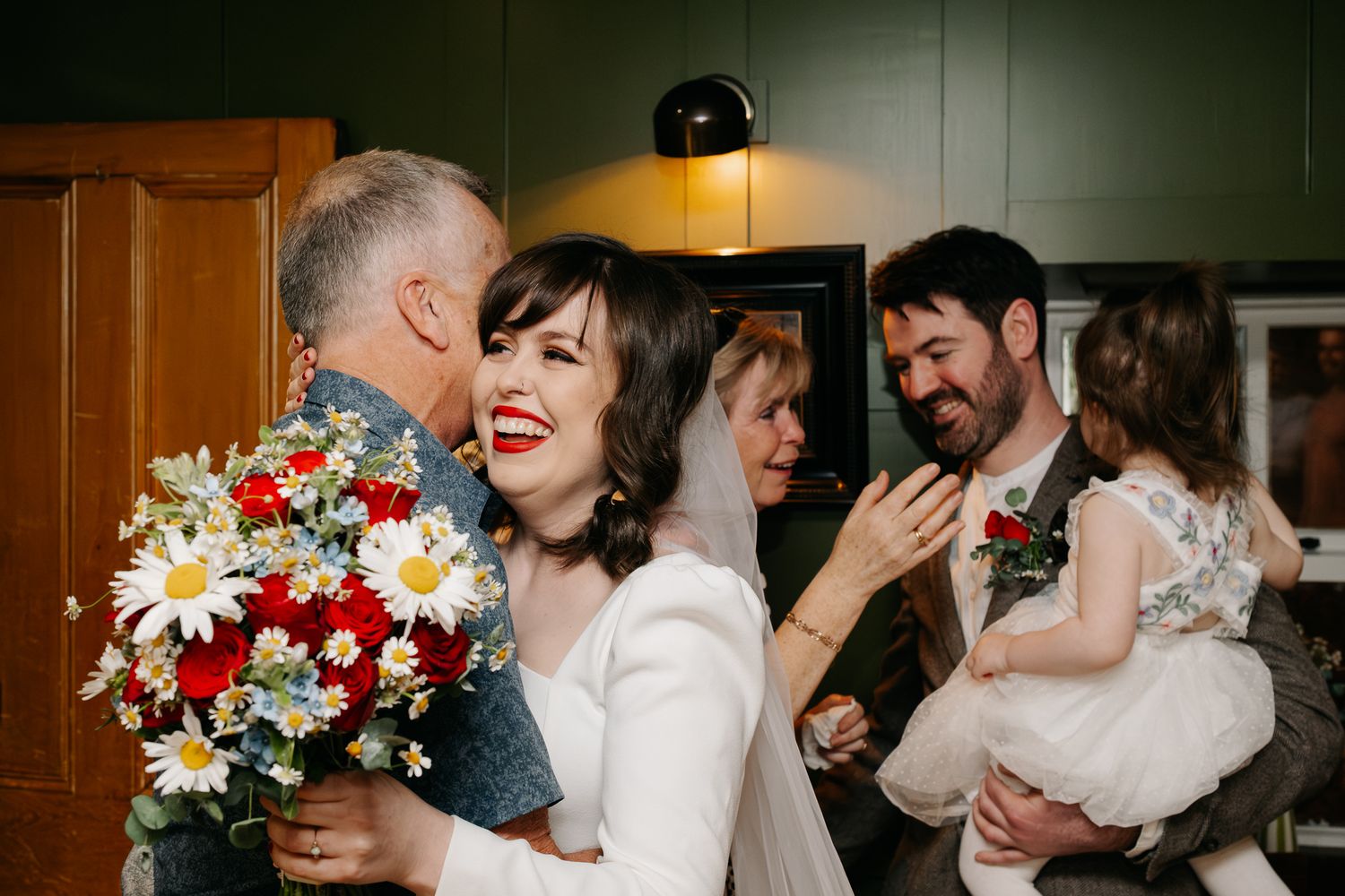 Bride with red lipstick holding colorful daisy bouquet smiles while guests celebrate at an indoor wedding reception.