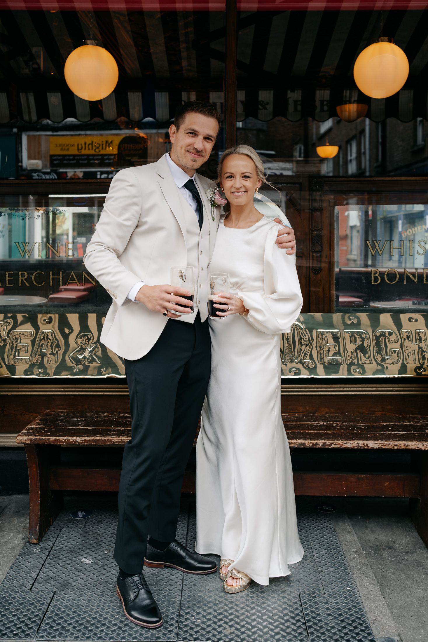 A smiling bride and groom in elegant wedding attire pose together outside a vintage pub, holding drinks.
