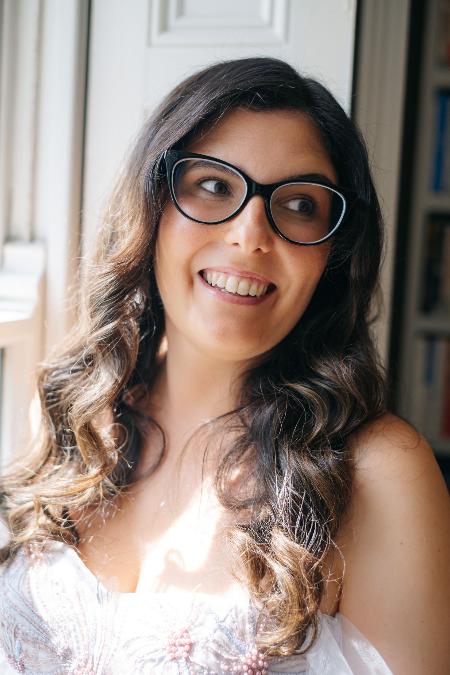Smiling young woman with curly dark hair and black cat-eye glasses wearing a white dress near a bright window.