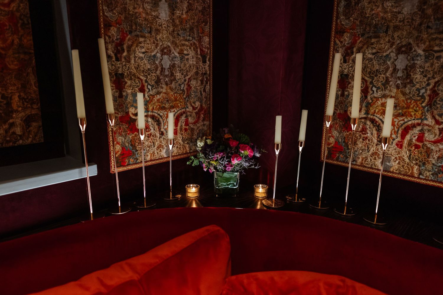 Elegant white candles in tall holders arranged on red velvet furniture against a patterned wall.