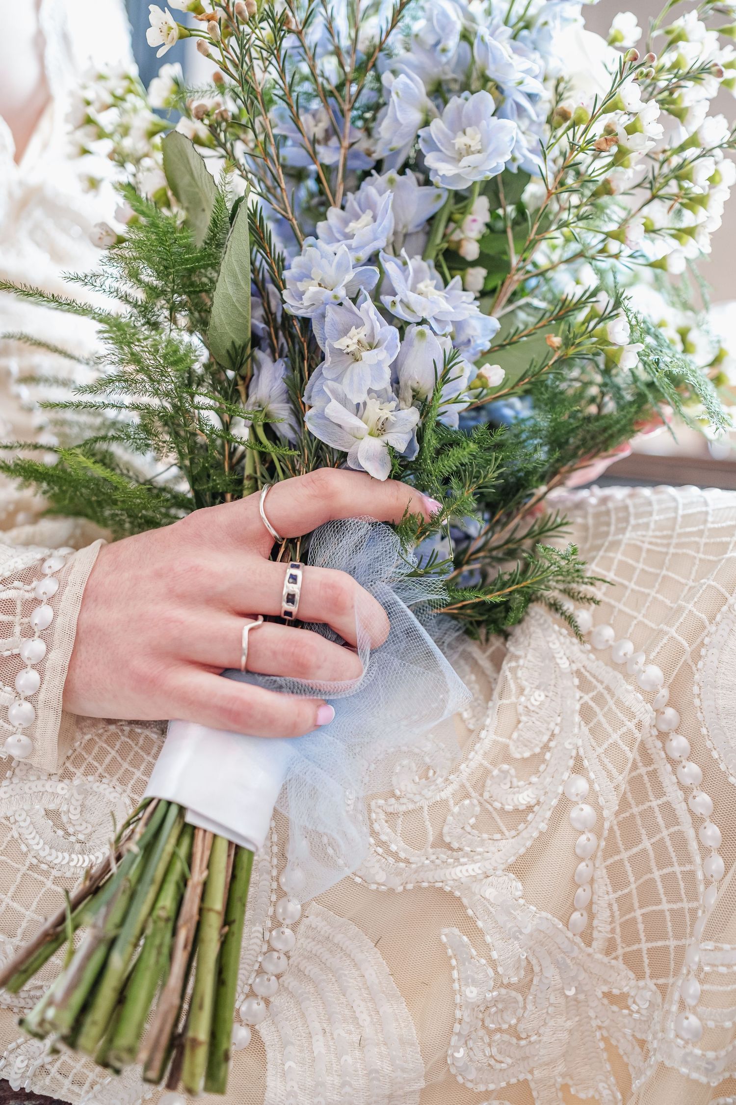 Close up of a delicate white lace wedding dress with the hands holding a bouquet of blue and white flowers.
