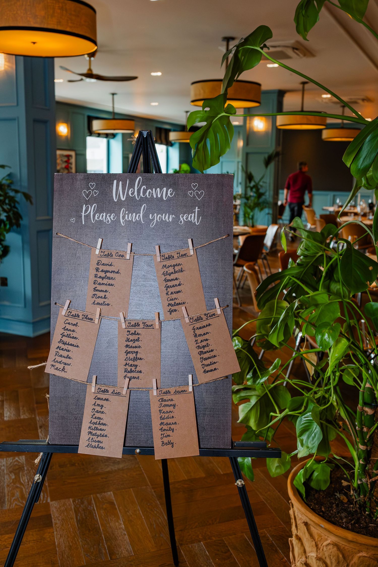 Rustic wooden seating chart display board with 'Welcome' text at a wedding reception venue with warm lighting.
