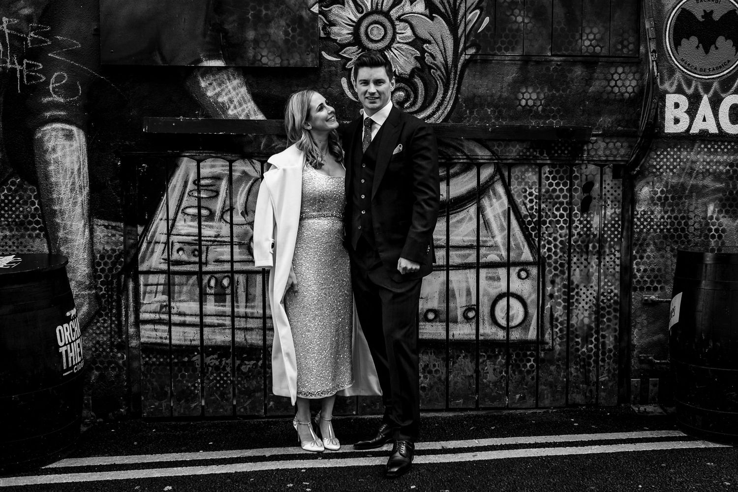 Wedding couple in formal attire pose against urban graffiti wall in black and white.
