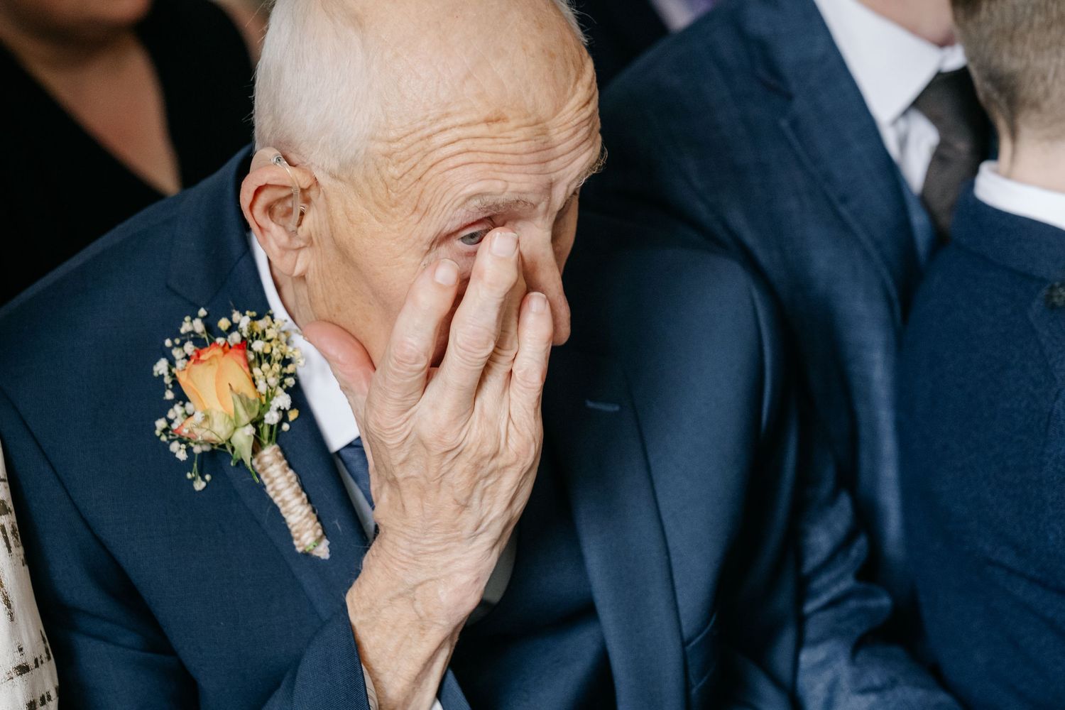 An emotional moment captured as someone wearing a navy suit and floral boutonniere covers their face during a wedding ceremony.