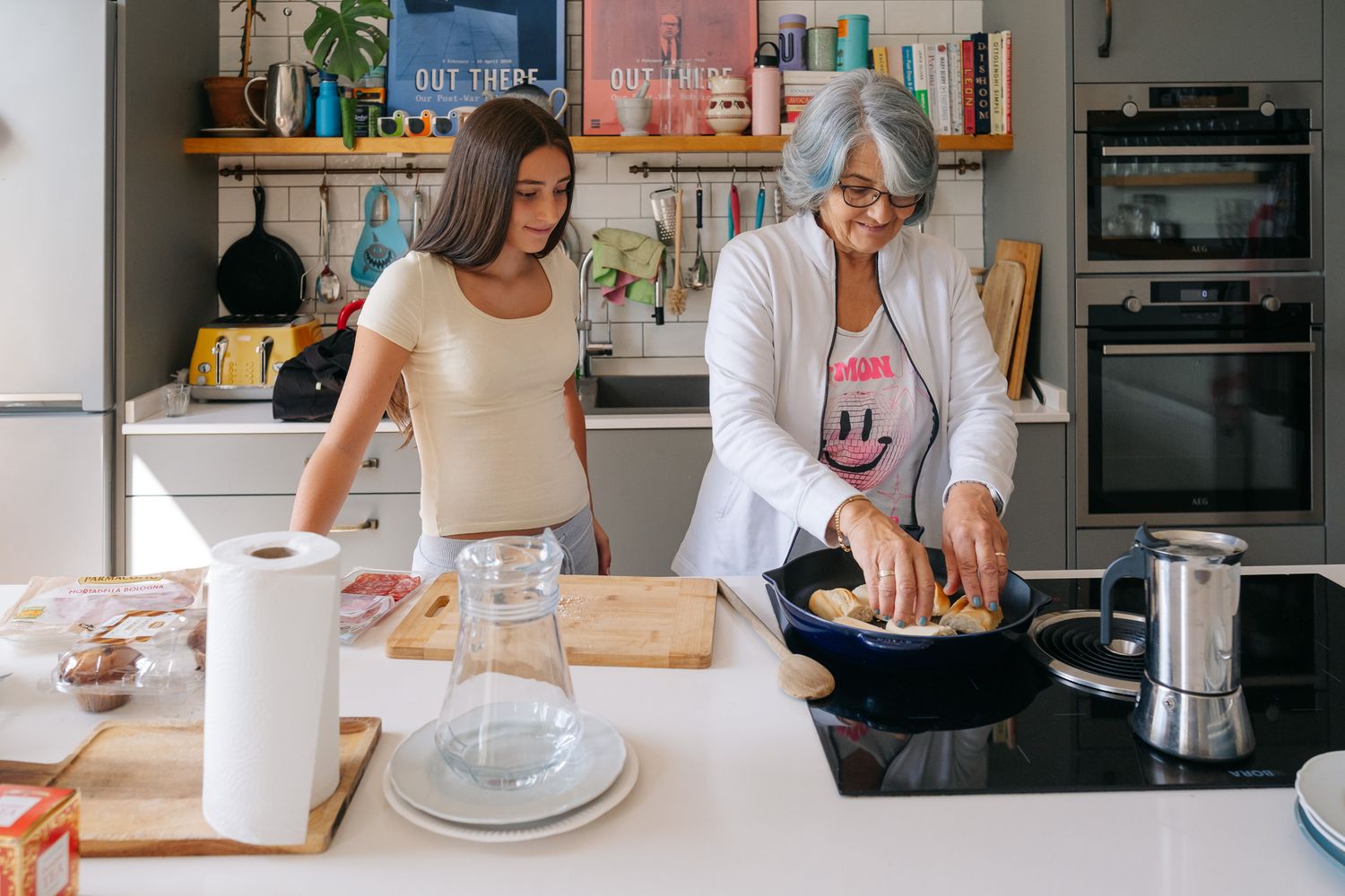 Older woman and teenage girl cooking together in a modern kitchen, stirring food in a pan on the stove.