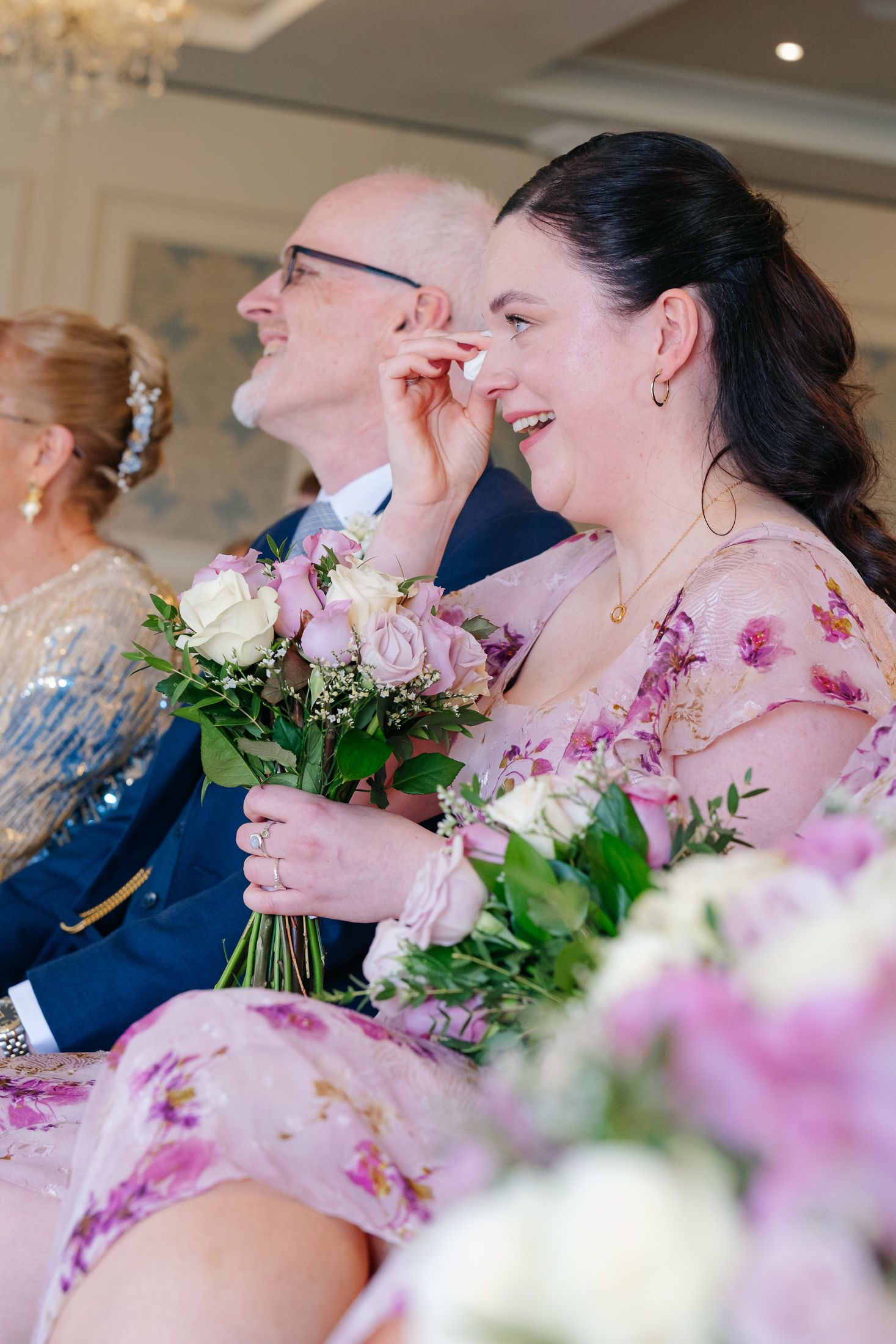 Emotional bridesmaid in floral dress wiping tears while holding pink rose bouquet during wedding ceremony.