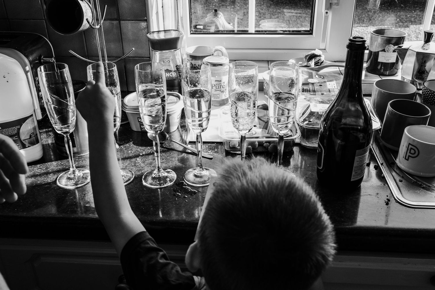 Black and white photo of a child reaching for champagne flutes on a kitchen counter with a wine bottle nearby.