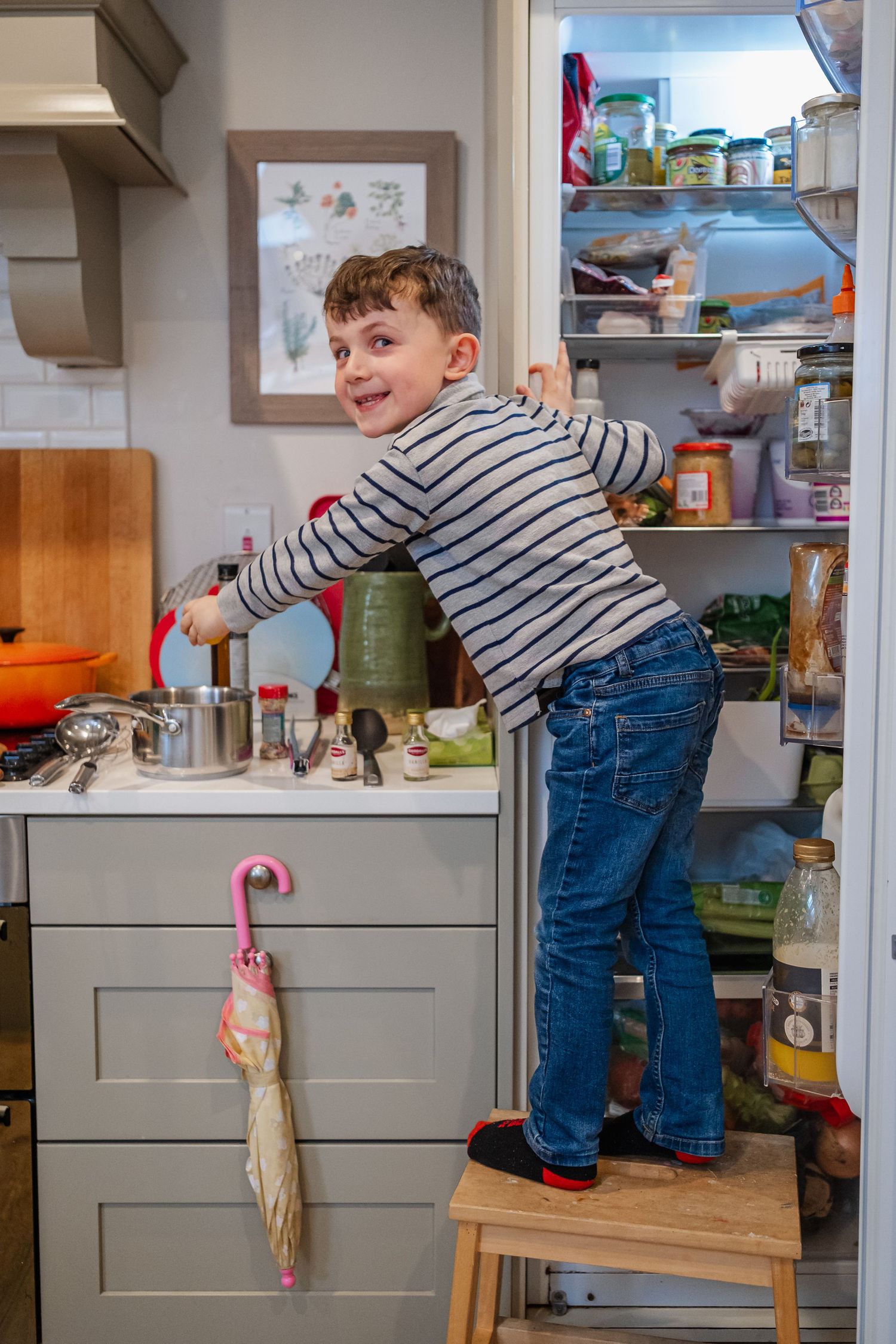Young boy standing on a stool reaching into an open refrigerator in a modern kitchen wearing striped shirt and jeans.