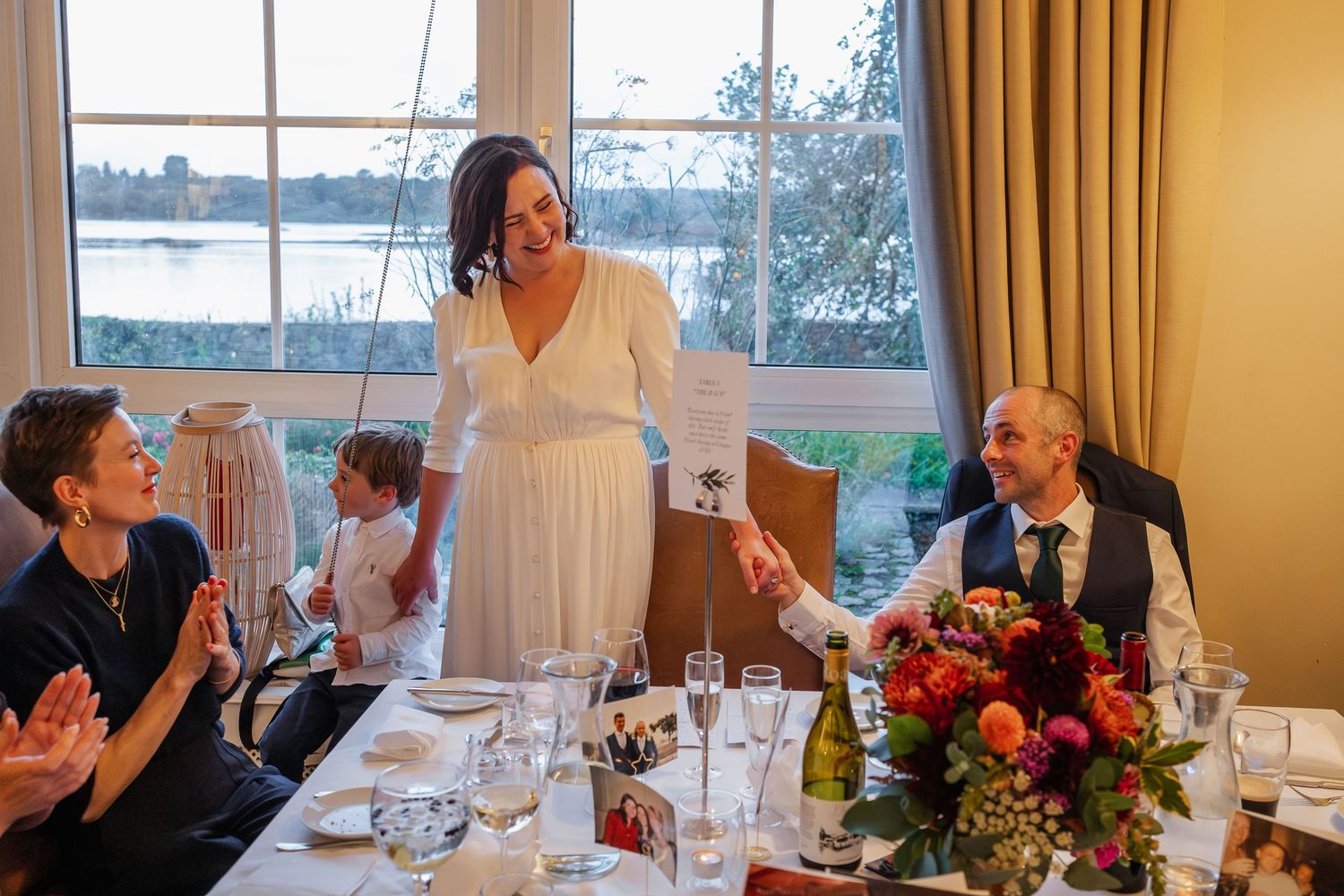 Bride in white dress standing and speaking at wedding reception table with waterfront view.