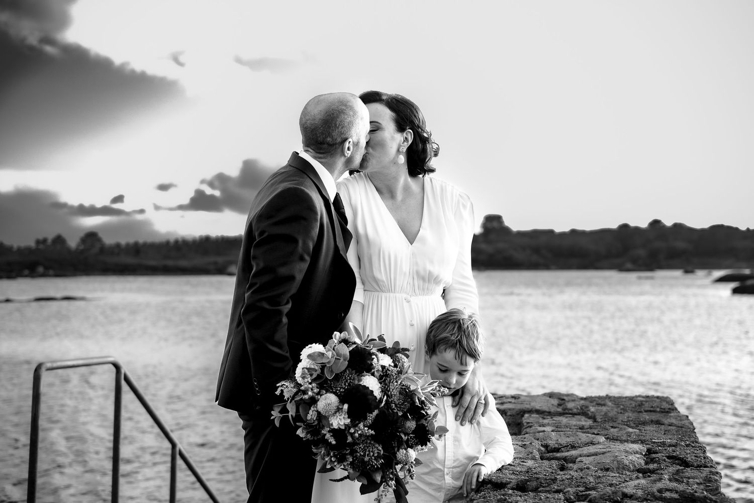 Black and white wedding photo of couple kissing by waterfront at sunset, bride holding bouquet, child standing nearby.