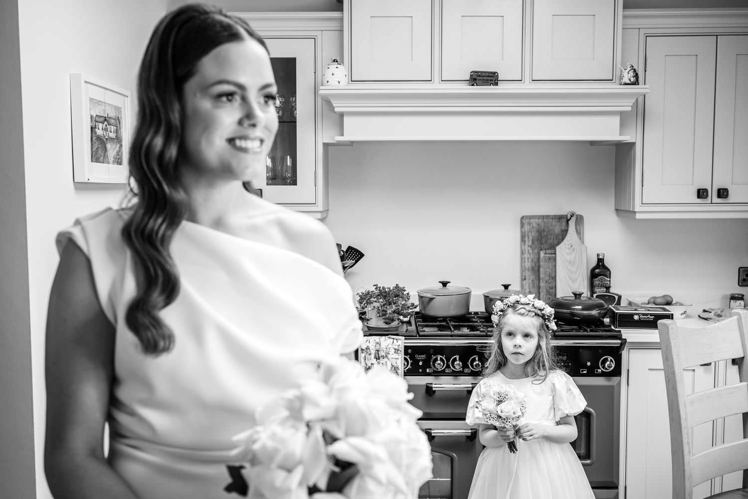 Black and white photo of bride and flower girl standing in kitchen on wedding day.
