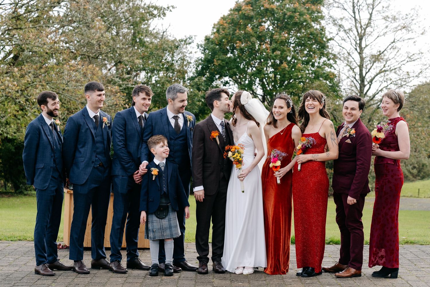 Wedding party in navy suits and burgundy dresses pose outdoors in a garden setting.