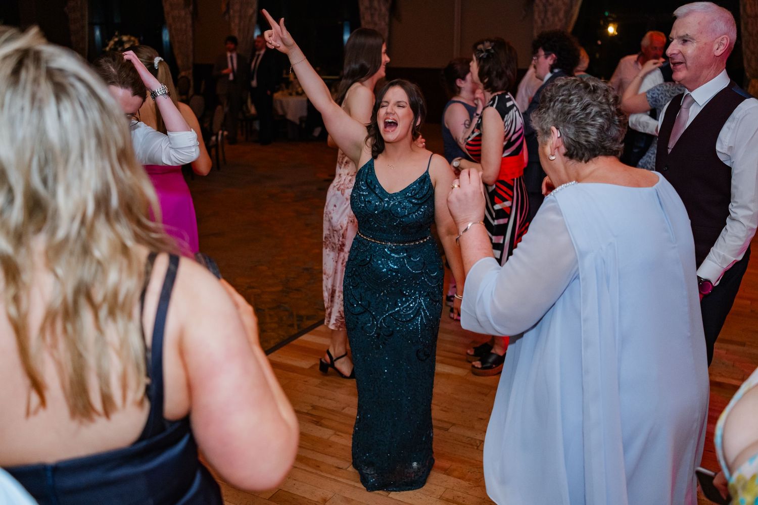 Wedding guests dancing together on a wooden dance floor at an indoor reception venue.