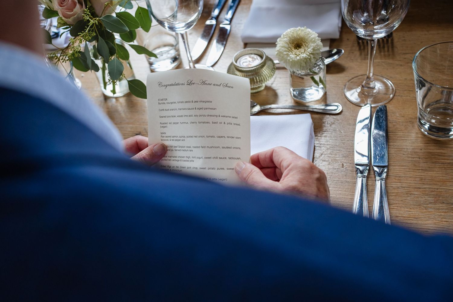 Someone holds a white menu card while seated at an elegant table setting with glassware and floral centerpieces.