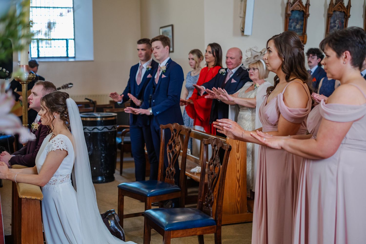 Wedding ceremony attendees in various pastel dresses observe from wooden church pews during a special moment.